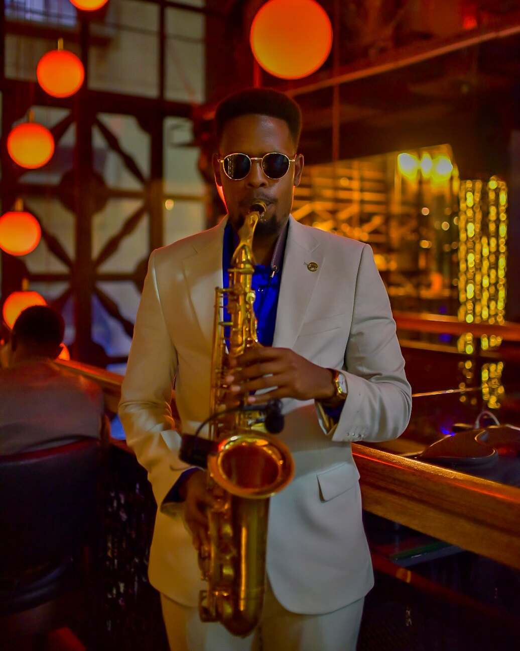 Ian in a cream jacket and blue shirt playing saxophone in a lounge lit by orange globe lamps