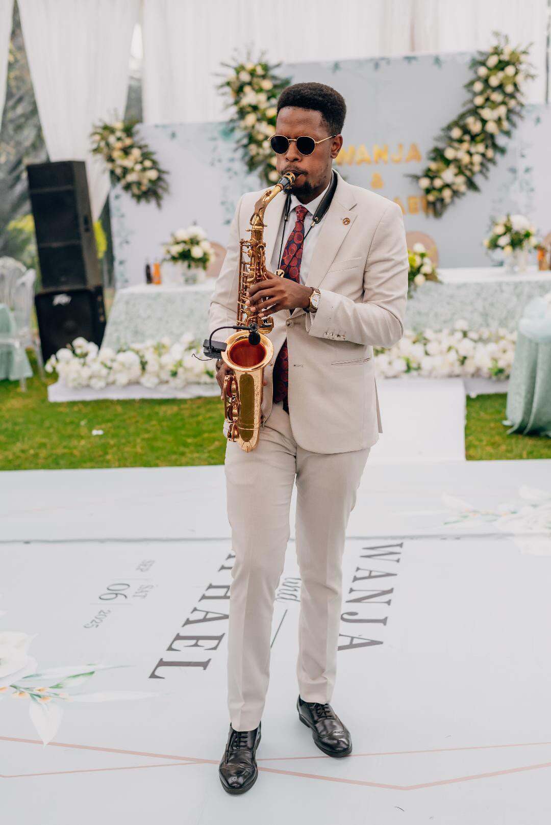 Ian in a beige suit and round sunglasses playing saxophone across a personalised white dance floor at a garden wedding
